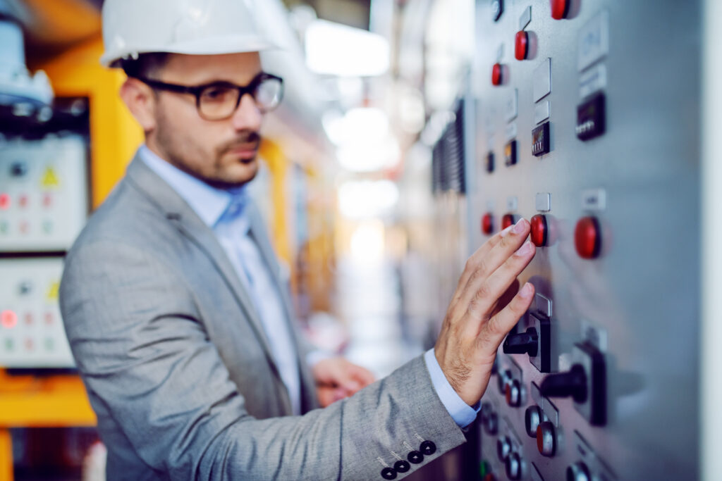 Serious handsome caucasian supervisor in gray suit and with helmet on head turning switch on. Selective focus on hand. Power plant interior.